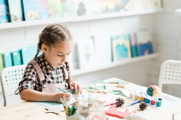 Cute casual schoolgirl with paintbrush drawing Christmas picture by desk while working individually