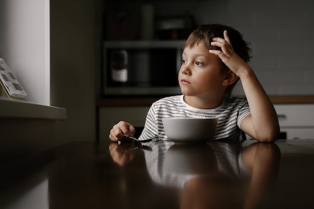 Cute caucasian 6 year old boy eating oatmeals for breakfast and looking in window