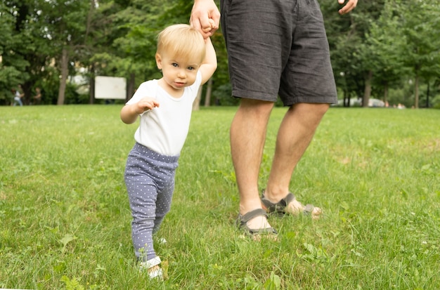 Bambino biondo caucasico sveglio in piedi sull'erba che tiene la mano dell'uomo irriconoscibile, che guarda l'obbiettivo. papà e bambino che camminano nel prato. genitore e figlio, trascorrere del tempo insieme all'aperto.