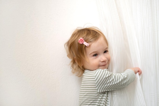 Cute caucasian blonde baby girl of 12 year old playing with white curtainshaving fun at home in light interior looking at cameraKid childtoddler games indoorslaughingsmiling infantCopy space