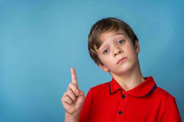 A cute Caucasian boy of 5 years old in a red T-shirt thoughtfully, seriously and edifiingly points his finger up. Blue background. copy space