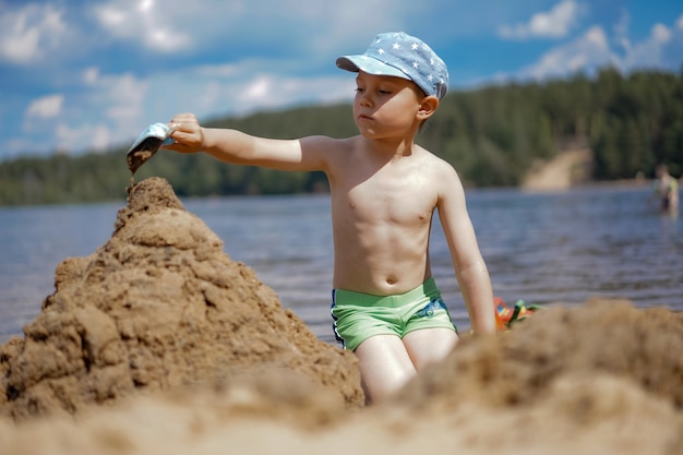 Cute Caucasian boy making sand castle on the beach of forest lake pouring water from the shovel to