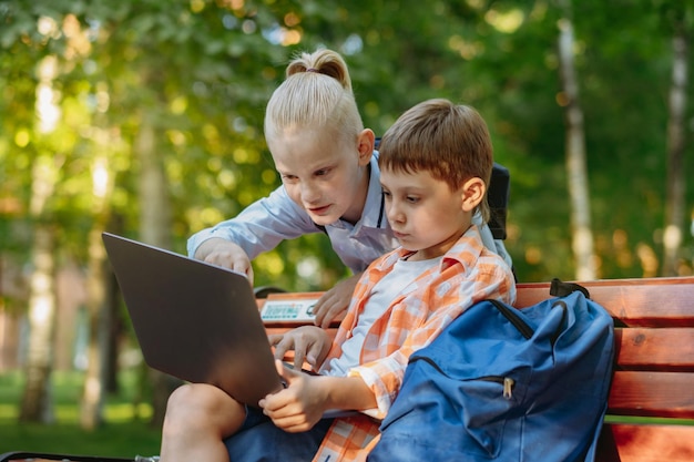 ラップトップ コンピューターで公園のベンチに座っているかわいい白人の男の子。学校の後に屋外で宿題をやっています。