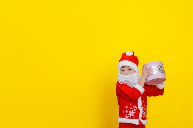 Cute Caucasian child boy in Santa Claus clothes and with a beard raised a pink round gift box