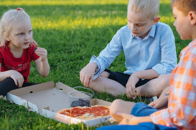 Cute caucasian children sitting on grass in park having lunch eating pizza outdoors after school