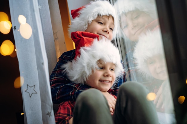 Cute caucasian children wearing snta hats and plaid shirts sitting hugging by the window waitinf for christmas