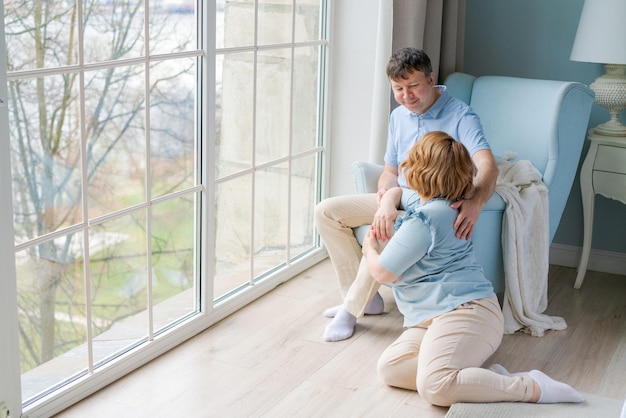 Cute caucasian couple relaxing at home man sits in an armchair and woman on floor next to him by a large window Enjoy serene happiness in a bright bedroom in casual clothes