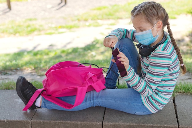 Photo a cute caucasian girl in a mask sits in the schoolyard with a backpack and a phone in her hands