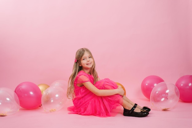 Cute Caucasian girl of seven years old in a bright pink barbicore style dress sits on a pink background with festive water balloons for her birthday