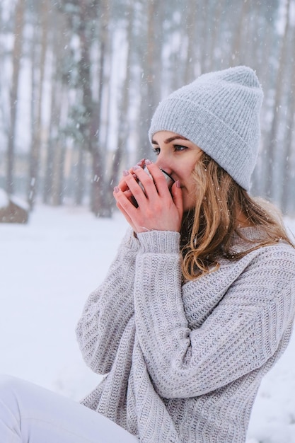 Cute Caucasian girl in a white sweater and hat in a snowy forest Park drinking hot tea or chocolate from a thermos. Girl Wearing Warm Winter Clothes during Christmas holidays, New Year