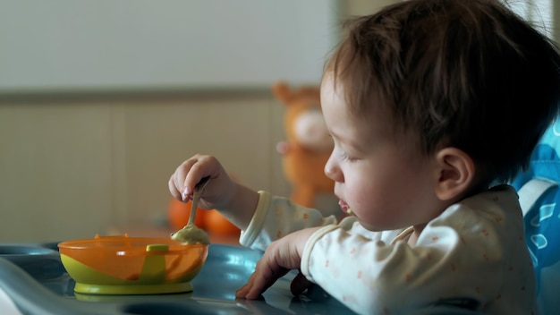 Cute Caucasian kid boy sitting in high chair eating broccoli puree with spoon