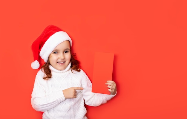 Cute caucasian kid girl in red santa\'s hat points with his\
finger on certificate or greeting card empty copy space for design\
or text banner christmas mockup template