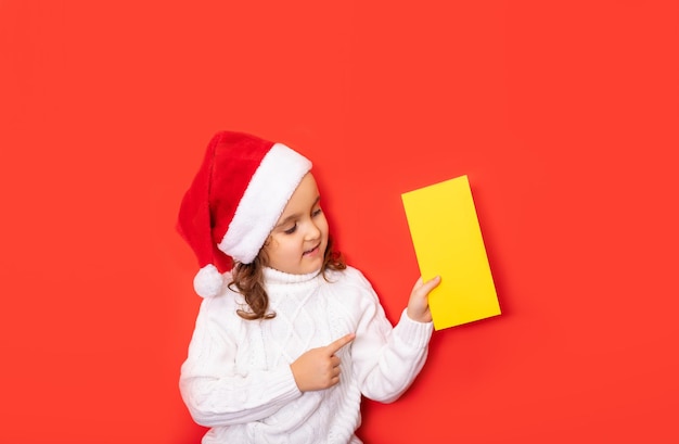 Cute caucasian kid girl in red santa\'s hat points with his\
finger on certificate or greeting card empty copy space for design\
or text banner christmas mockup template