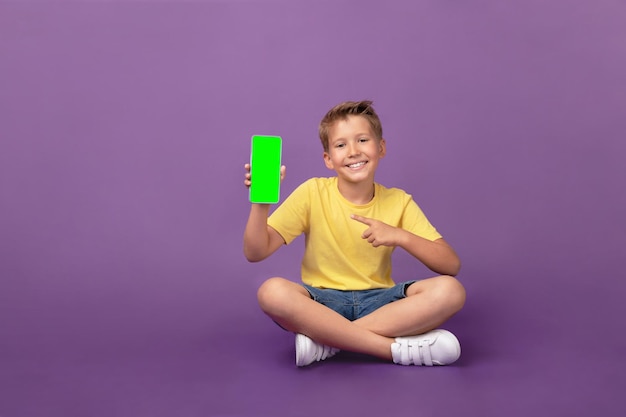 Cute Caucasian little boy pointing at mobile phone phone with chroma key green screen and looking at camera Smiling child with cell phone on purple background Studio shot