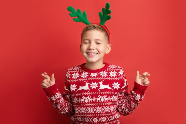 Cute Caucasian little boy wearing Christmas reindeer antlers and sweater makes wish for christmas with closed eyes. Happy child staying crossed fingers isolated on red. Studio shot.