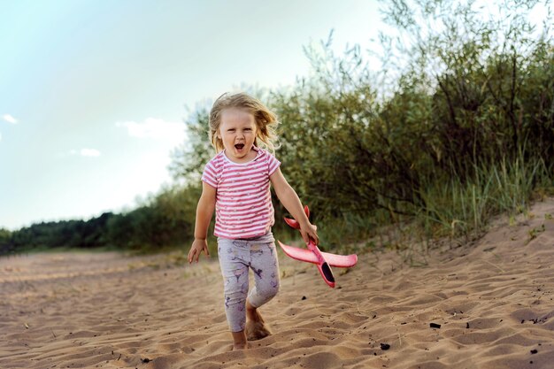 Bambina caucasica carina che corre lungo la spiaggia per lanciare un aereo giocattolo