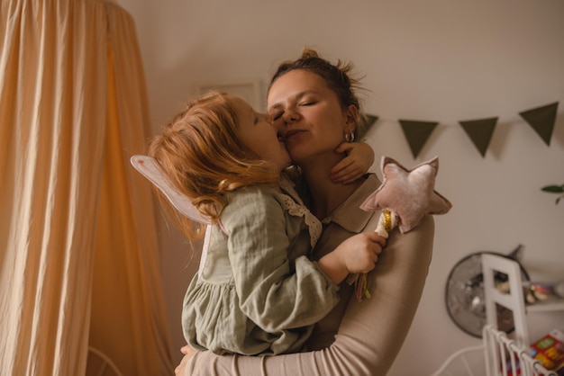 Cute caucasian redhaired girl kisses her mother wear casual clothes on her arms while standing in children's room Family love concept