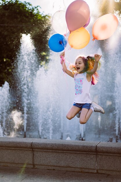 cute caucasian smiling girl with colorful dyed hair holding baloons jumping from fountain