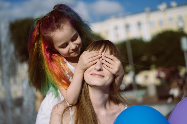 cute caucasian smiling girl with colorful dyed hair standing behind mom closing her eyes holding bal