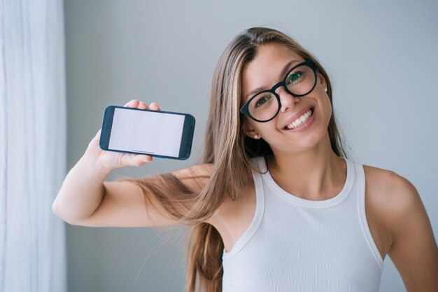 Cute caucasian young woman in glasses and white tshirt holds\
phone shows blank screen looks at camera broad smiles stands at\
home pretty female student satisfied by phone app technology