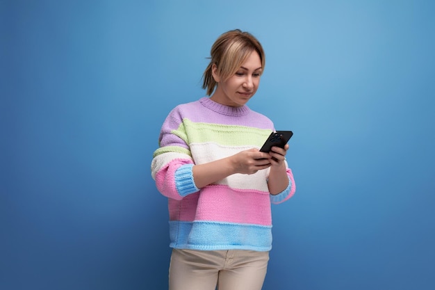 Carina giovane donna bionda affascinante in un maglione a strisce che chiacchiera in uno smartphone su sfondo blu