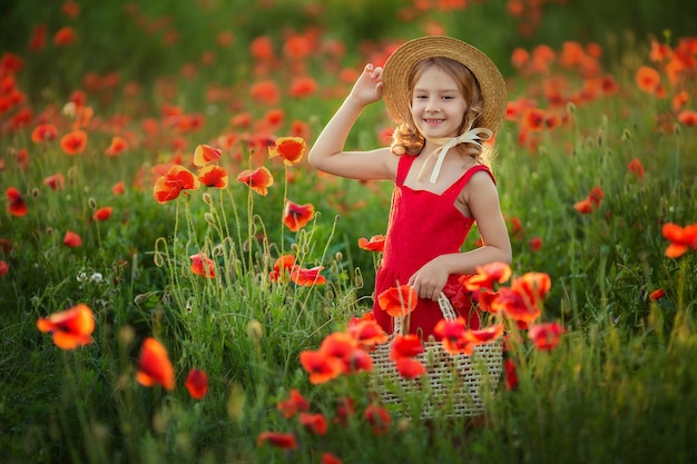 cute charming girl with blond hair in a straw hat and a red dress holds a basket with poppies in a poppy field and sniffs flowers