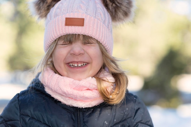 ピンクのニット帽と手袋をしたキュートで陽気な青い目のブロンドの女の子は、冬の雪の上で公園で遊ぶ