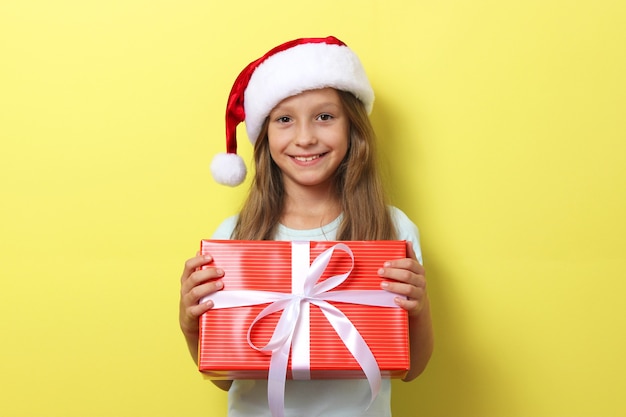 Cute cheerful girl in a christmas hat on a colored background holding a gift