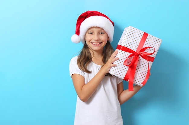 Cute cheerful girl in a christmas hat on a colored background holding a gift