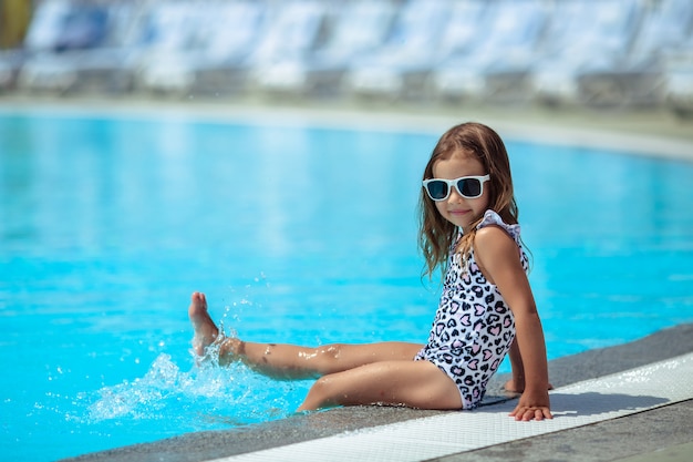 Cute cheerful girl in glasses and swimwear sits by the pool and splashes her legs in the water.