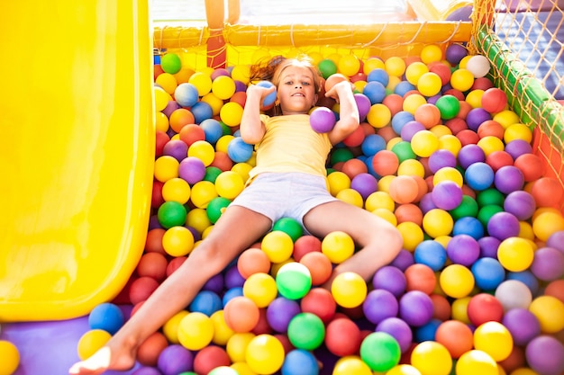 Photo a cute cheerful girl lies on the playground with soft and bright equipment and throws colorful balloons into the camera, enjoying the warm summer sun