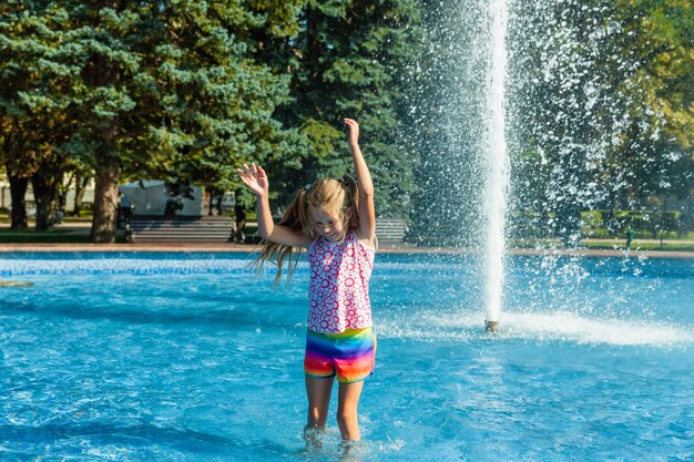 귀여운 명랑 소녀가 분수에서 놀고 있습니다. 아이는 도시 분수에 있는 여름 공원에서 즐거운 시간을 보내고 있습니다.