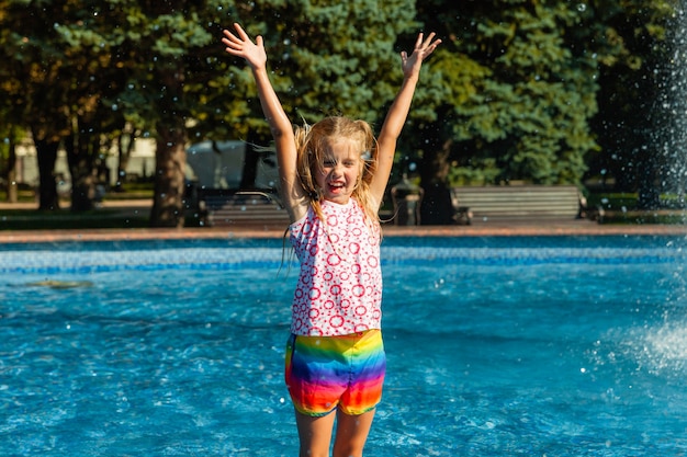 かわいい陽気な女の子が噴水で遊んでいます。子供は市の噴水のある夏の公園で楽しんでいます。