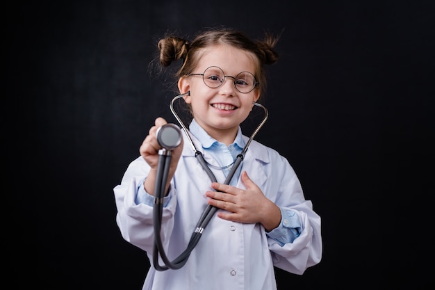 Милая веселая маленькая девочка в белом халате, держащая стетоскоп перед камерой, стоя против черного пространства