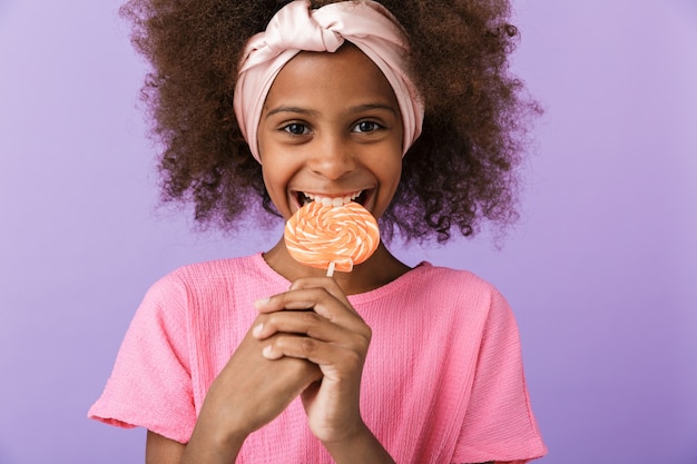 Carino allegro ottimista giovane ragazza africana ragazzo in posa isolato sul muro viola, mangiare caramelle lecca-lecca.