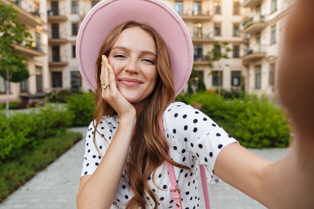 cute cheery young redhead woman walking outdoors by street in dress take selfie