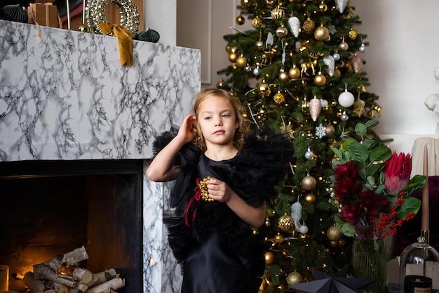 Foto bambina carina in abito nero con regalo di natale nelle sue mani vicino ad alberi di natale con luci buon natale e buone feste