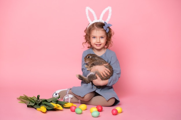 Cute child girl in bunny ears holding rabbit, tulips, colorful eggs.