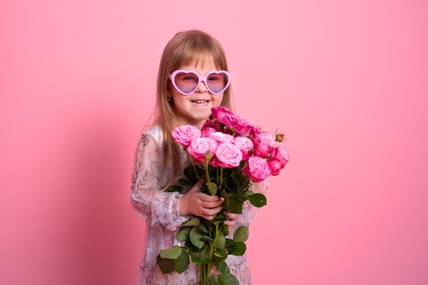 Cute child girl in dress sunglasses holding bouquet of pink roses on pink background