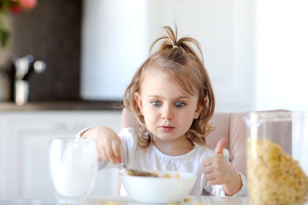 Cute child girl has breakfast and looks surprised to plate with corn flakes and milk