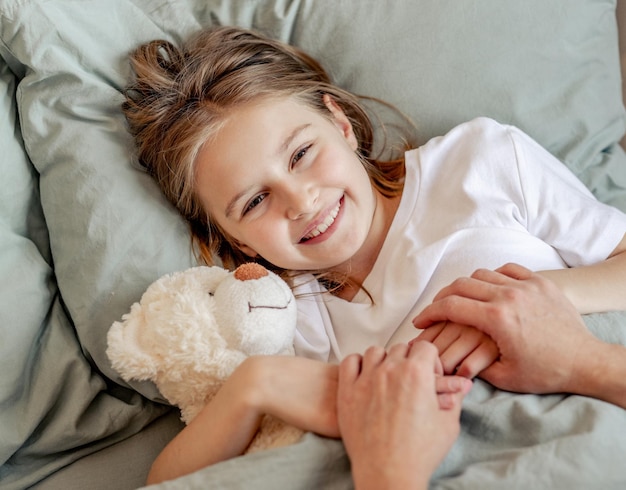 Cute child girl hugging teddy bear toy in the bed and looking at mother. Parent hands holding kid daughter in the morning