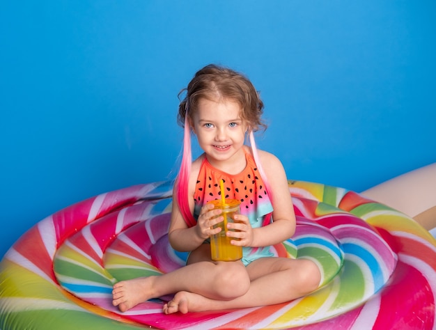 Cute child girl in swimming suit drinking orange juice sitting on colorful inflatable mattress on blue surface.