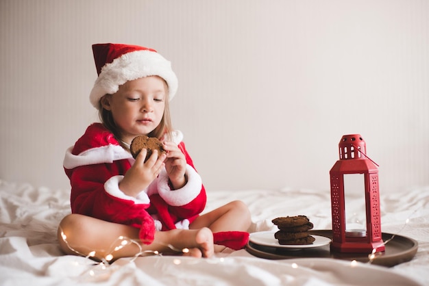 Милая девочка в красной шапке санта-клауса и халате сидит в постели с рождественским декором
