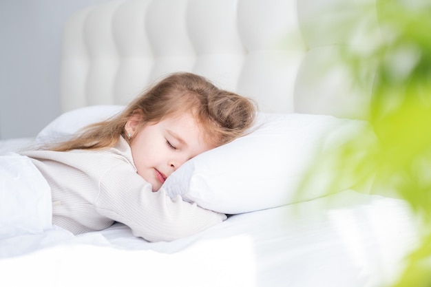 Cute child girl with long hair in beige pyjamas sleeping on white bedding at home