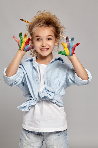 青いシャツと白いTシャツを着て、シックな巻き毛のブロンドの髪にブラシを持っているかわいい子供。彼女は灰色の背景に、彼女の塗られた腕で誰かを怖がらせています。