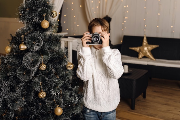 A cute child photographs on a retro camera against the background of an artificial Christmas tree with golden balls.