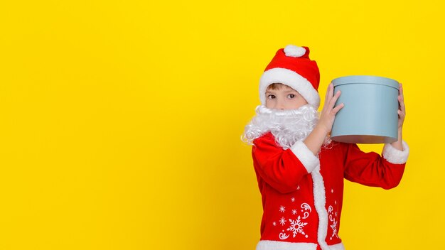 Cute child in Santa Claus clothes and with white beard holding a round gift box high in his hands