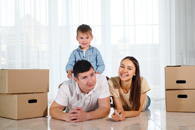 Cute child sits on father back lying on light floor in new house near beautiful wife among cardboard boxes