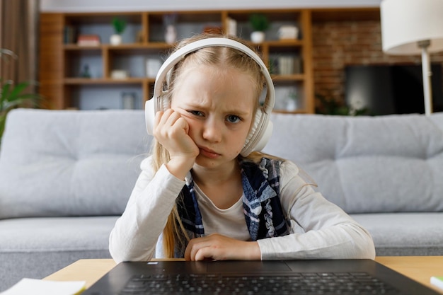 Cute child studying remotely at home using laptop tired primary\
schoolgirl falling asleep during an online lesson with teacher\
distance learning is boring and uninteresting web cam view