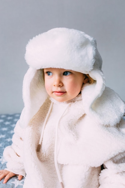 Cute child y on the bed in cozy adult clothes and a hat with earflaps